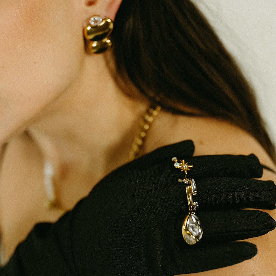 Close-up of a person wearing black gloves with gold jewelry, including earrings and a necklace.