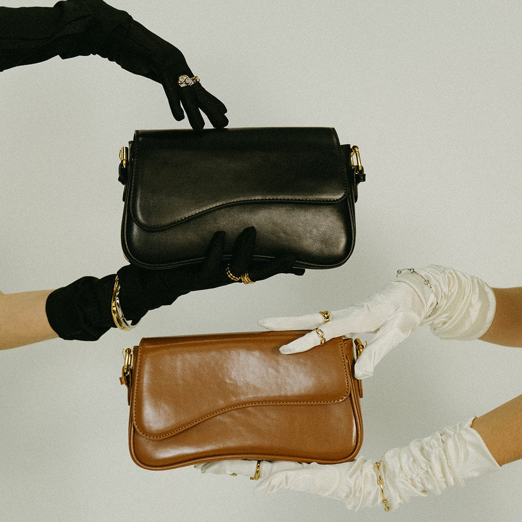 Two hands, one wearing a black glove and the other a white glove, holding a black and a brown leather handbag against a light background.