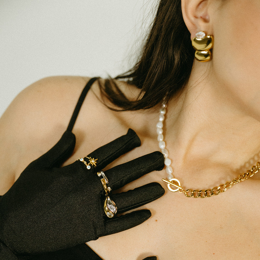 Person wearing black gloves, a pearl necklace, and gold jewelry with a neutral background